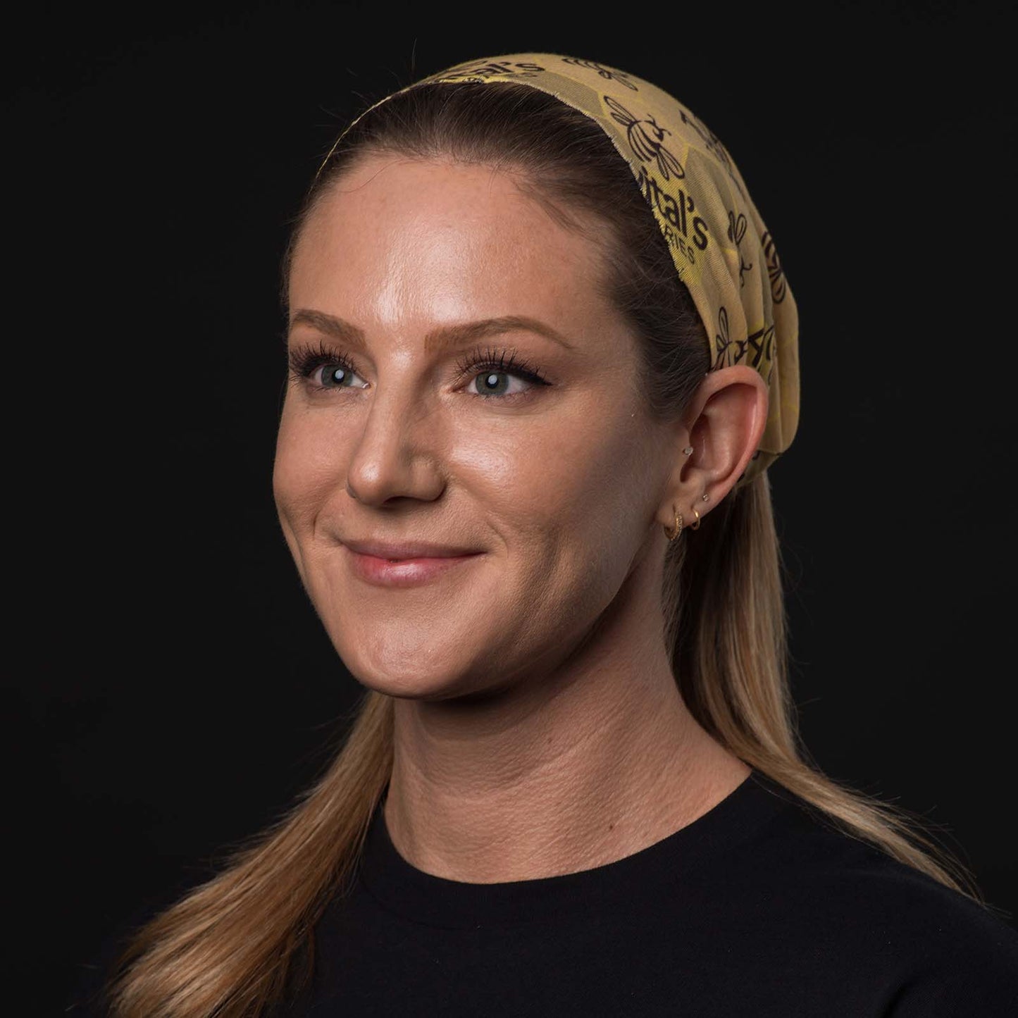 A blond woman smiling with an Avital's  tubular bandana holding her hair back. The bandana is yellow with a dark brown Avital's logo and honey bee stamped on it.
