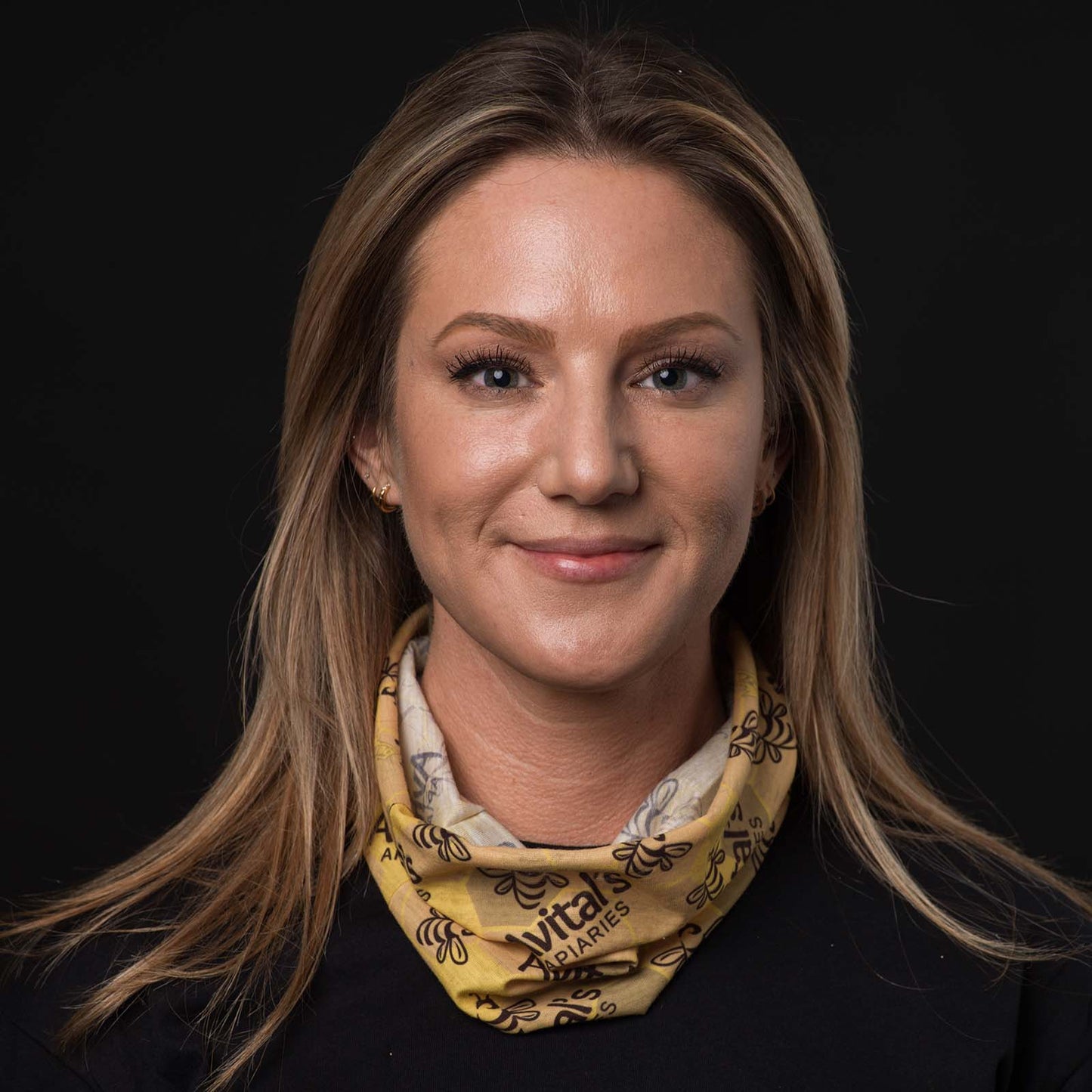 
                  
                    A blond woman smiling with an Avital's  tubular bandana around her neck. The bandana is yellow with a dark brown Avital's logo and honey bee stamped on it.
                  
                