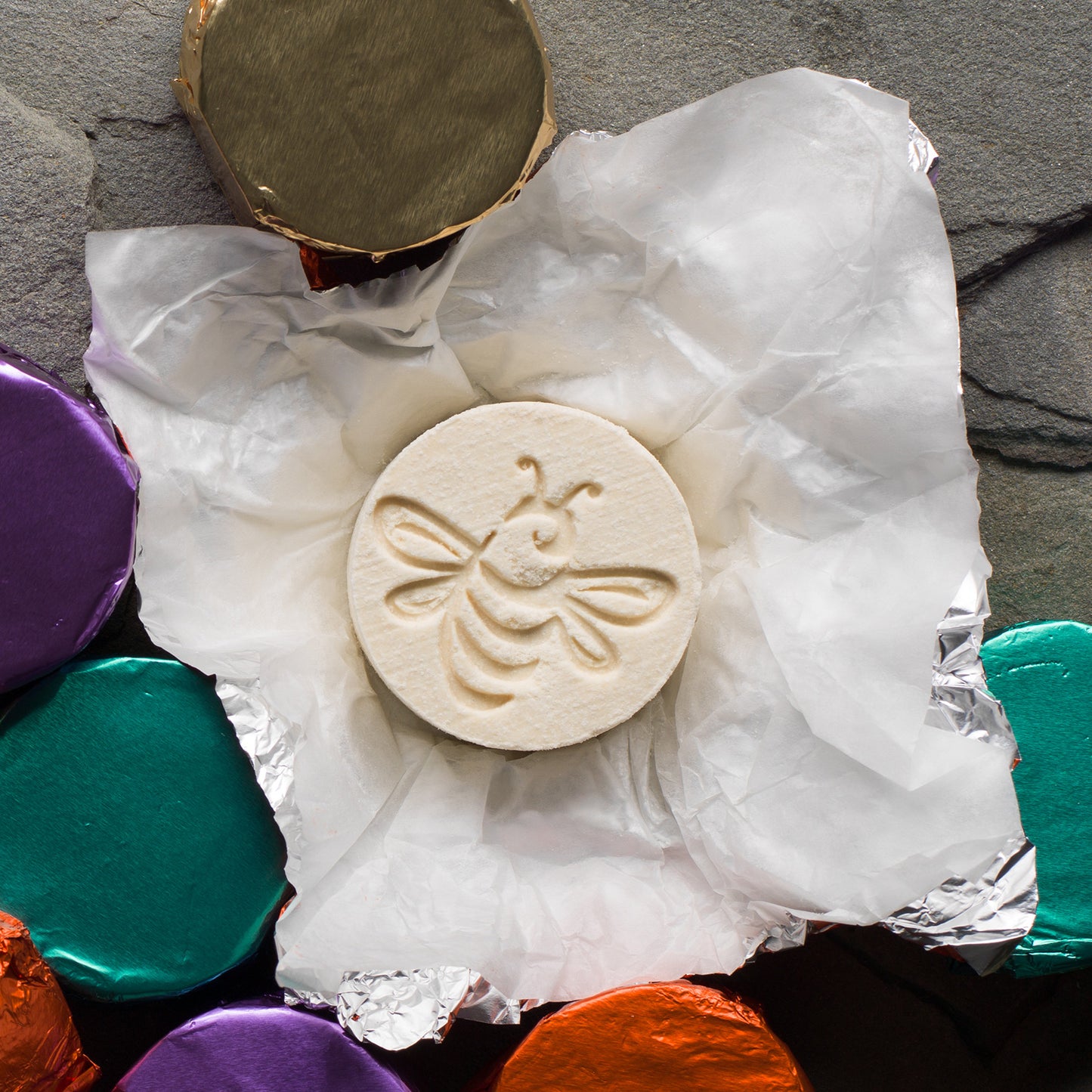 A bee-stamped bath fizzy on a tissue square surrounded by other fizzies wrapped in jewel toned foils.