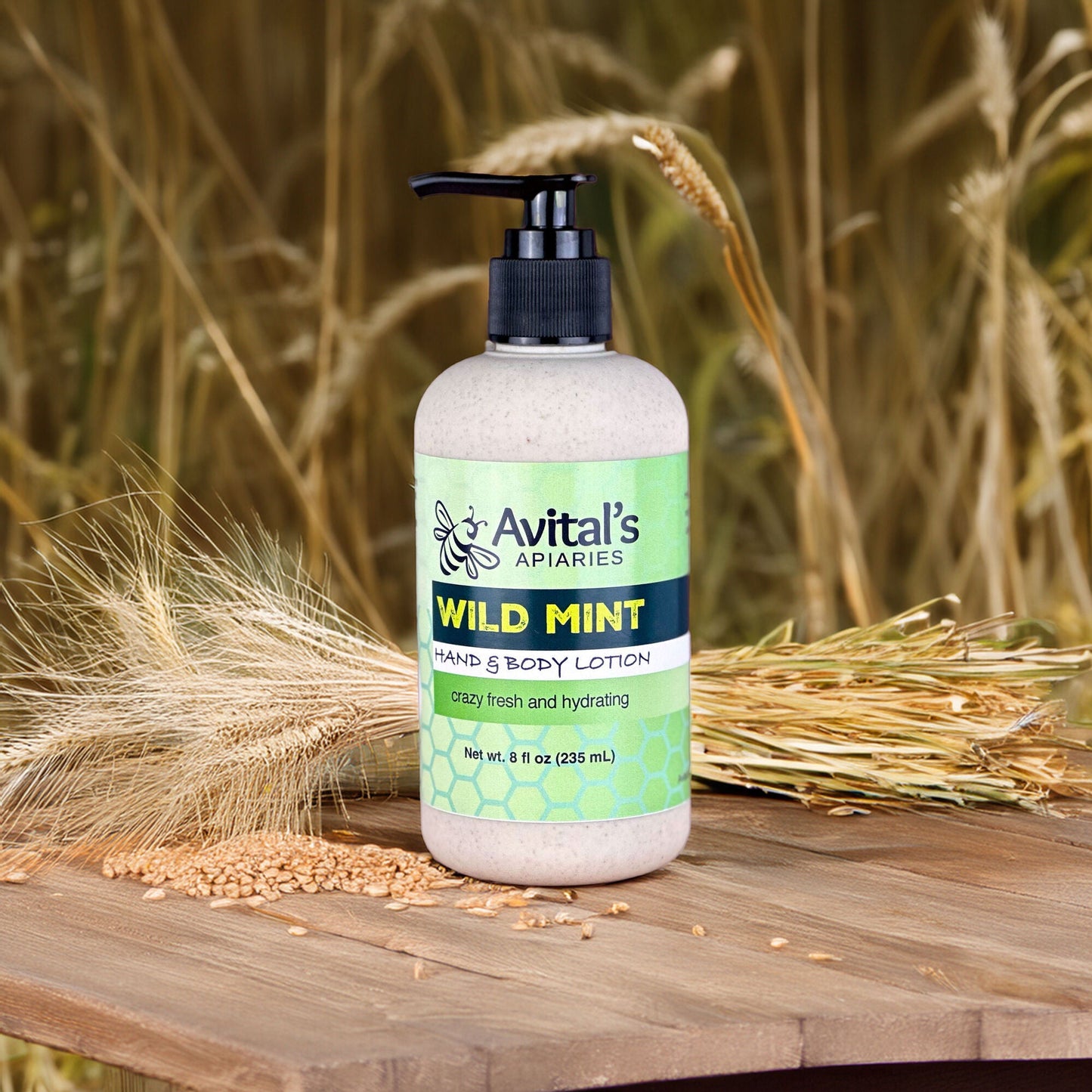 An 8 ounce straw-colored bottle with a black pump on a wooden table, surrounded by stalks of wheat and wheat kernels. The label is green and reads Wild Mint Hand & Body Lotion