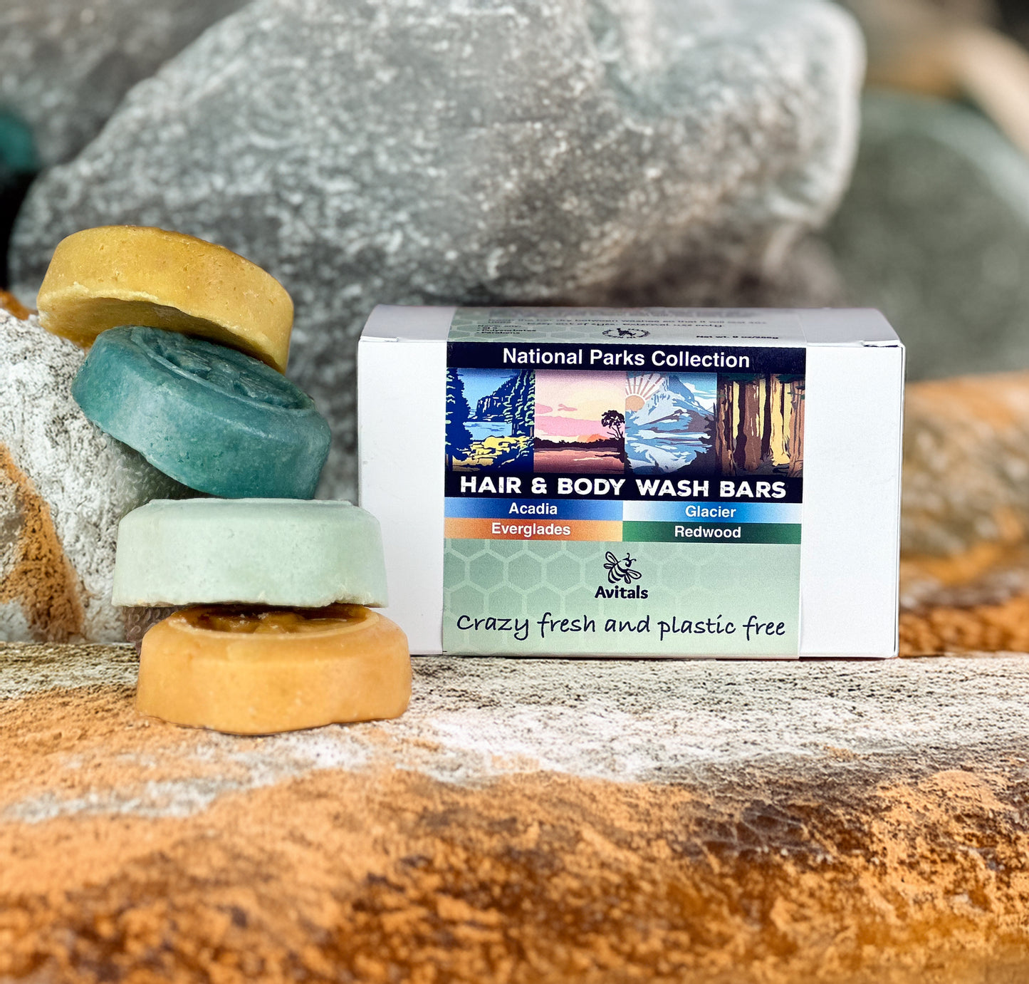A pack of four National Parks themed shampoo and body wash bars in various colors placed on a rock surface with a box labeled 'National Parks Collection Hair & Body Wash Bars'.