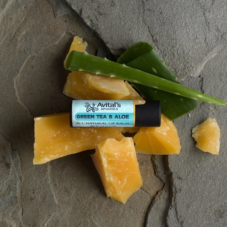A stick of lip balm labeled 'Green Tea & Aloe' in an eco-friendly paperboard tube, displayed on a natural stone background with pieces of beeswax and an aloe leaf.