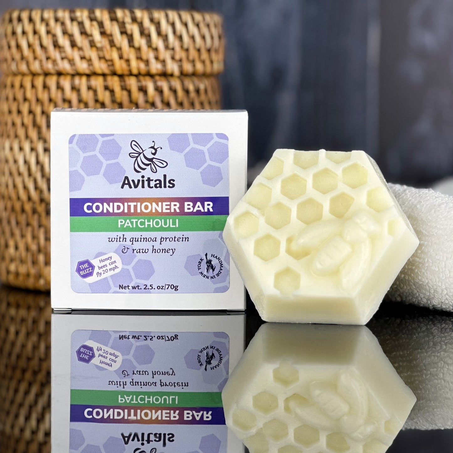 A Patchouli Conditioner Bar by Avital's Apiaries displayed alongside its packaging.   The bar is white and has a hexagonal shape, placed on a reflective surface with a wicker basket in the background.