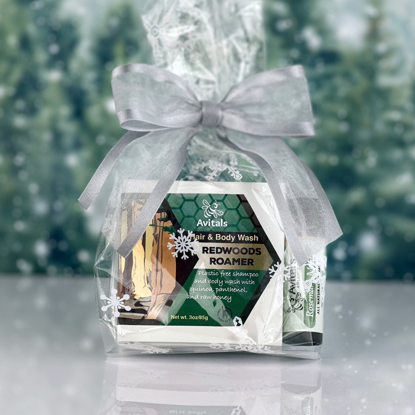 A gift pack containing a shampoo/body wash bar and a lip balm, both with 'Redwoods' and 'Rosemary Mint' labels, wrapped in a clear plastic bag with a white ribbon and snowflake decorations in the background.