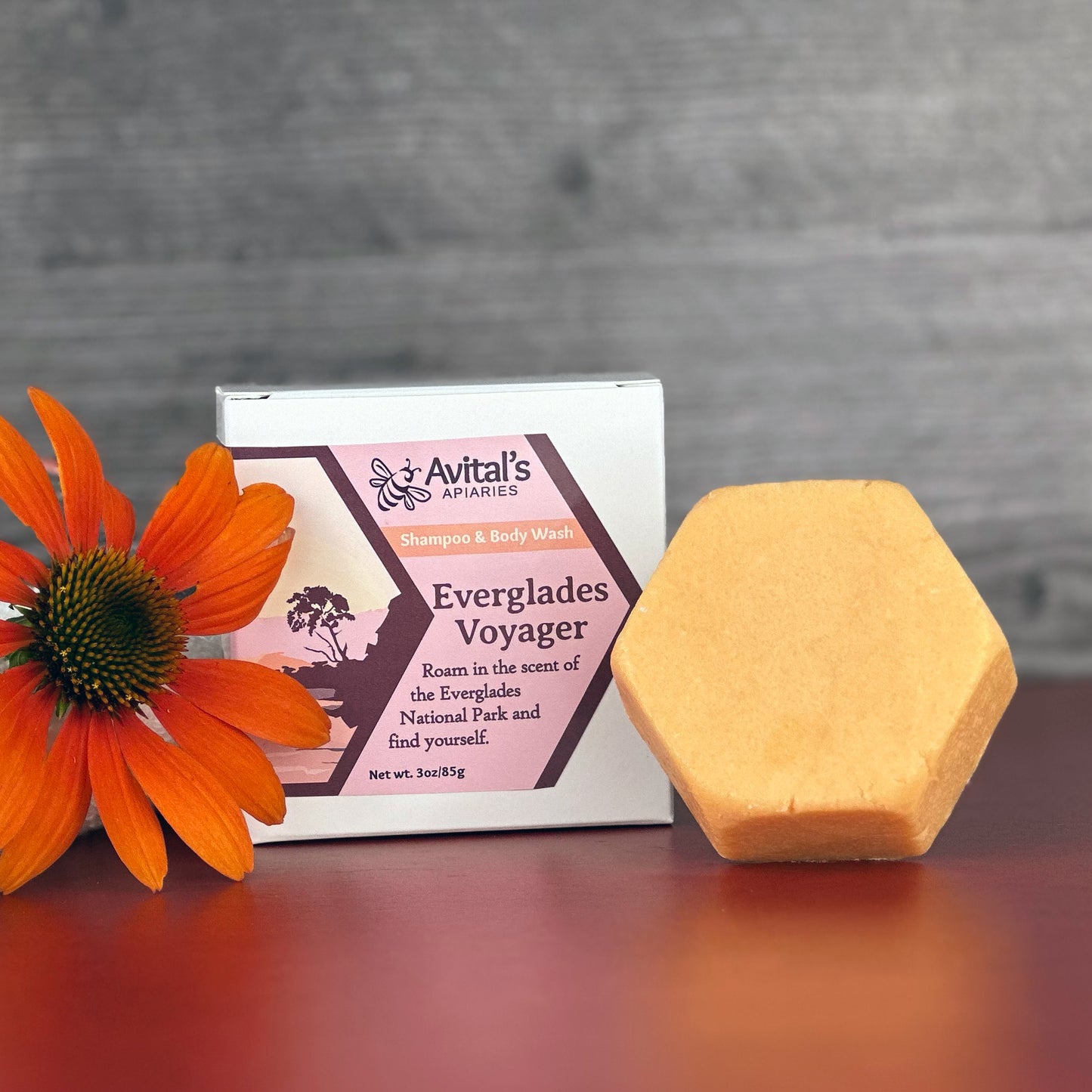 A bar of shampoo and body wash next to its packaging, labeled 'Everglades Voyager'. There is a flower with orange petals in the foreground to the left.