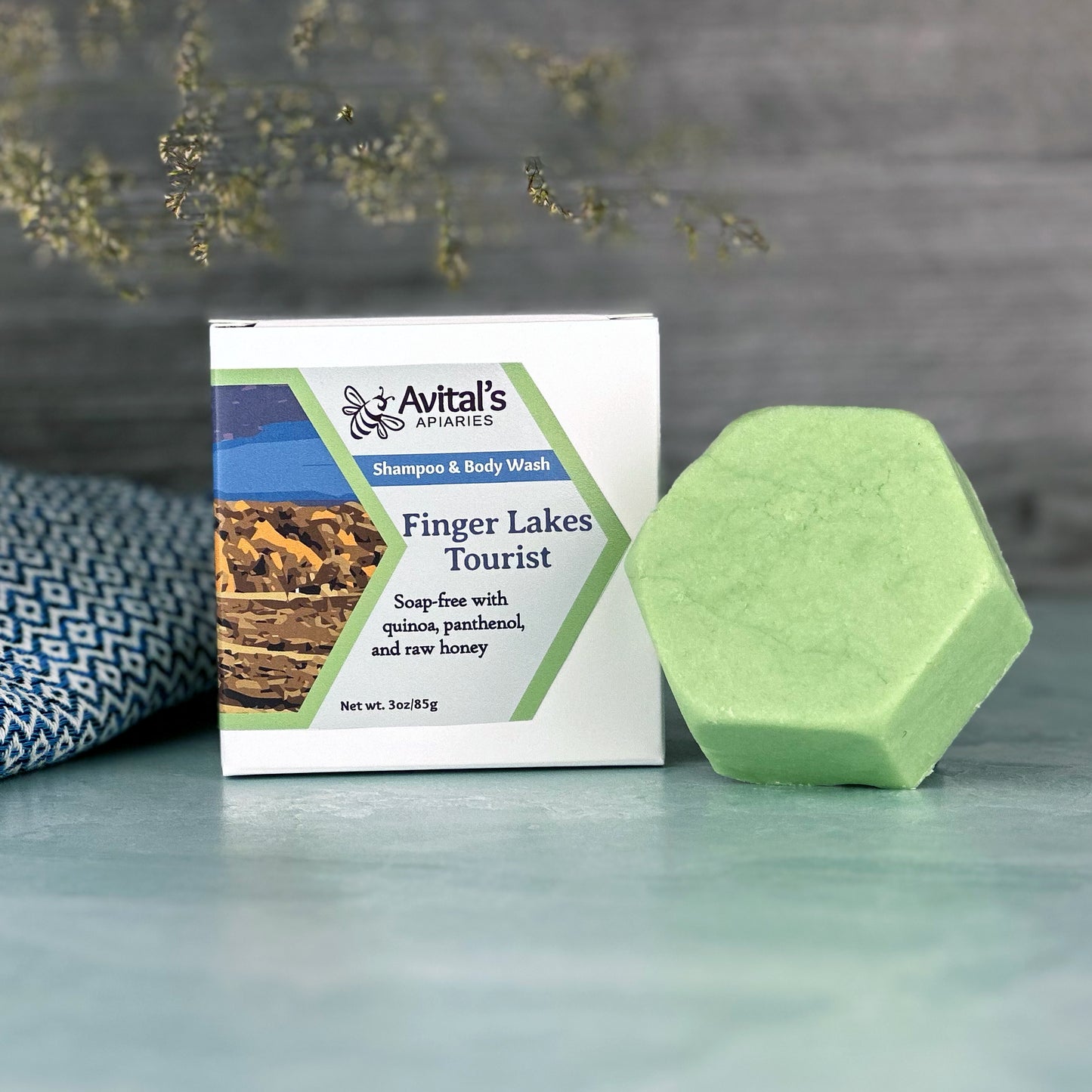 Green soap bar and packaging labeled 'Avital's' on a gray surface with a a towel and greenery