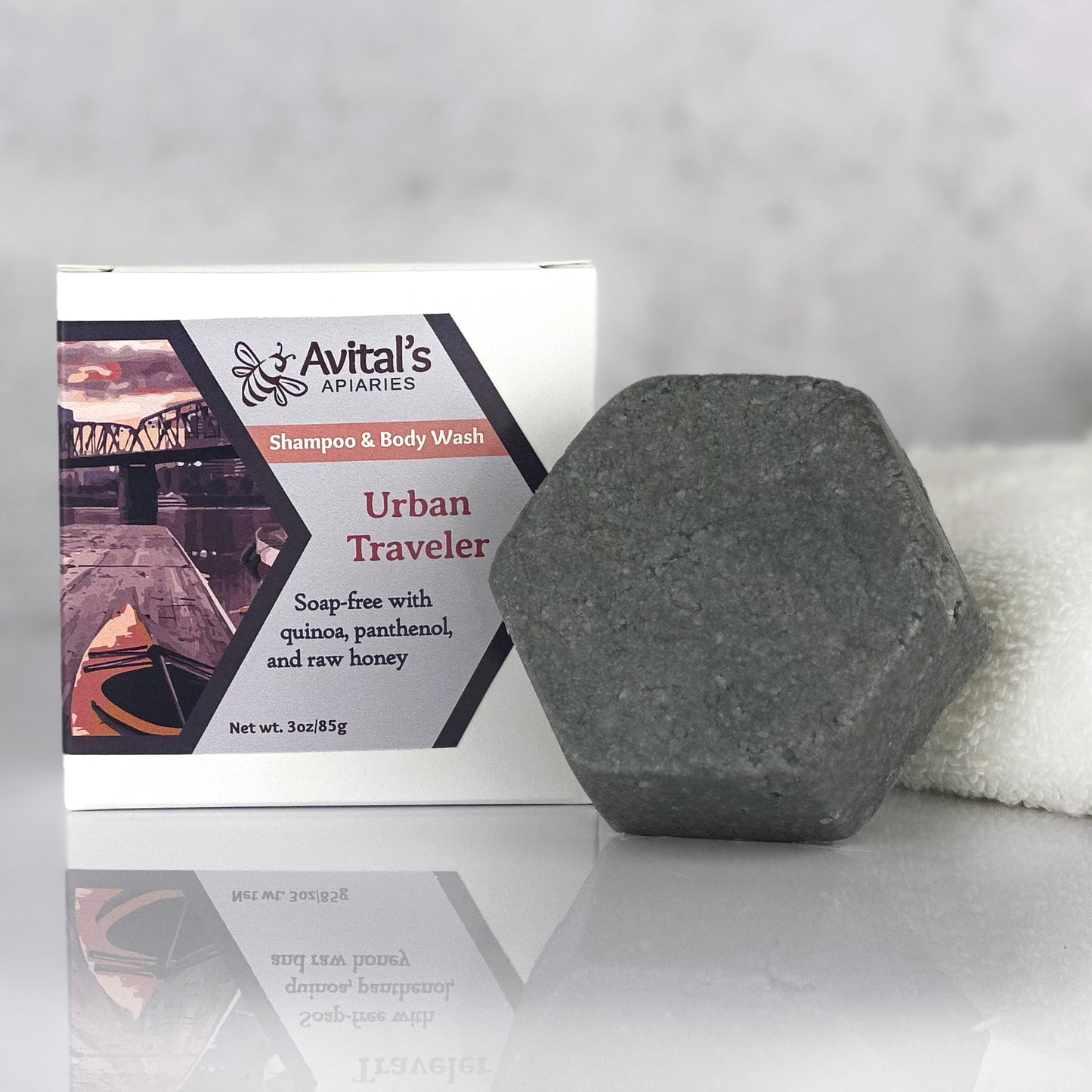 A dark gray hexagonal bar next to a bo: Avital's Apiaries Urban Traveler on a reflective surface with a white towel near by.