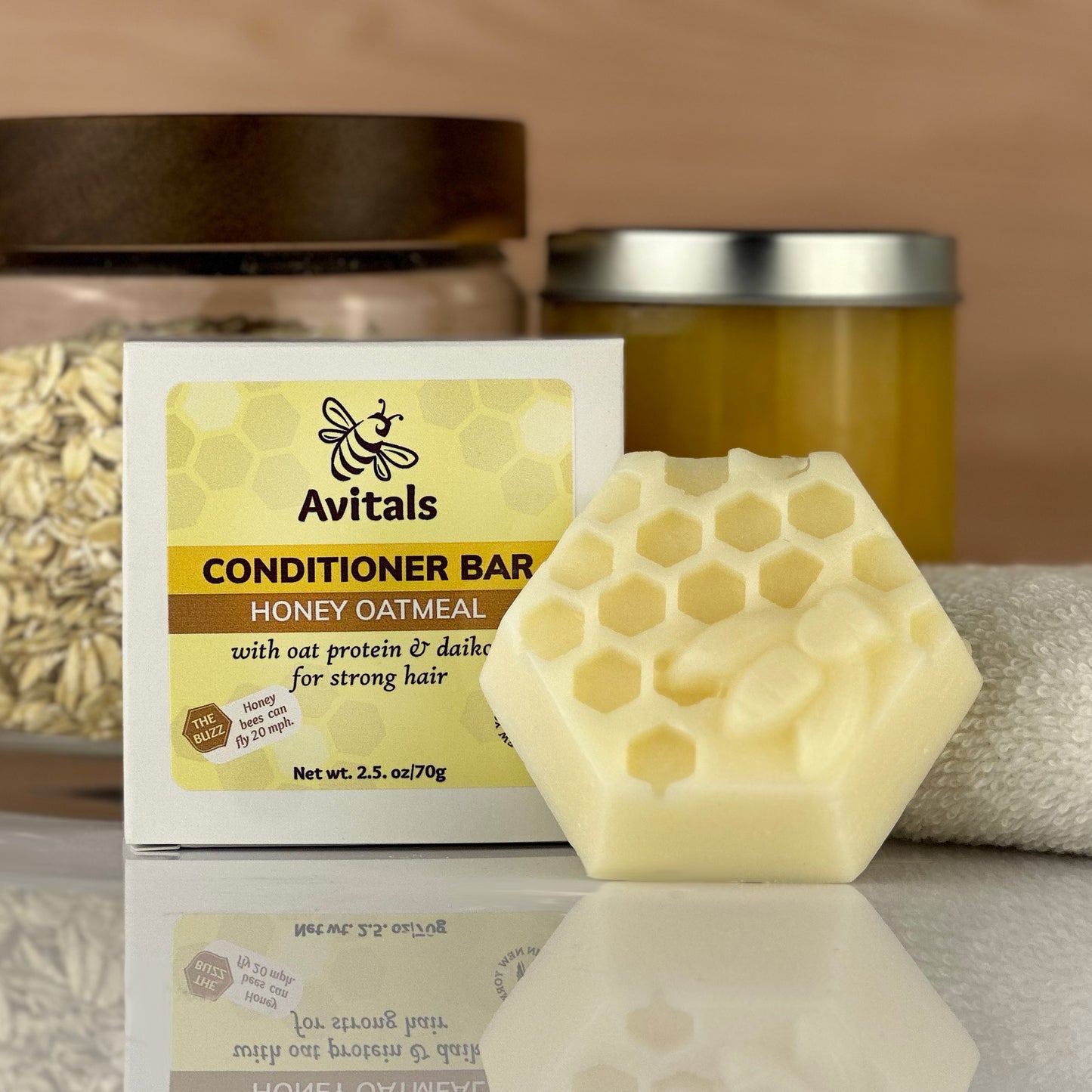 Avital's Honey Oatmeal Shampoo box and hexagonal bar on a reflective surface with a jar of oatmeal, honey, and a towel in the backgroun.