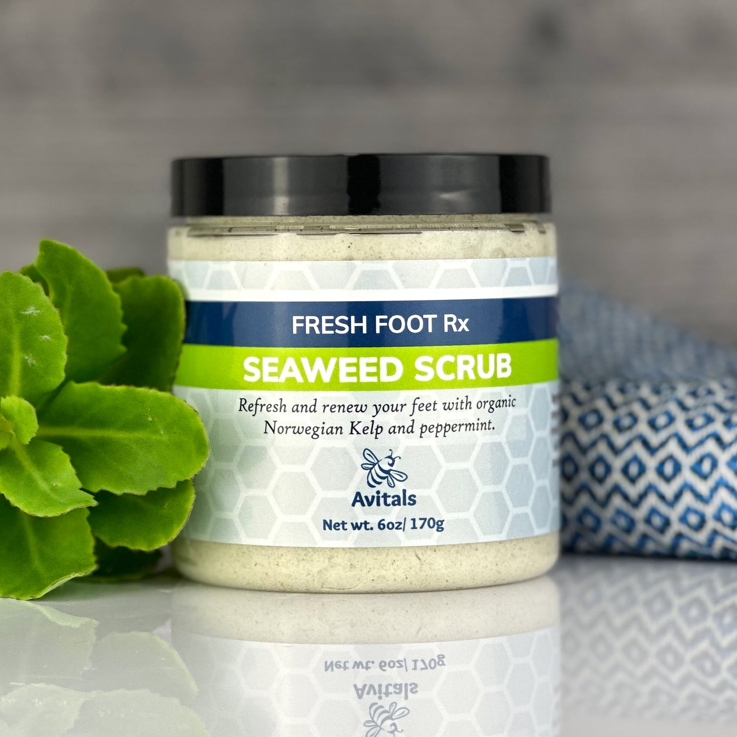 A jar of Fresh Foot Rx Seaweed Scrub by Avitals, with peppermint oil, placed on a surface alongside a sprig of peppermint and a patterned towel.