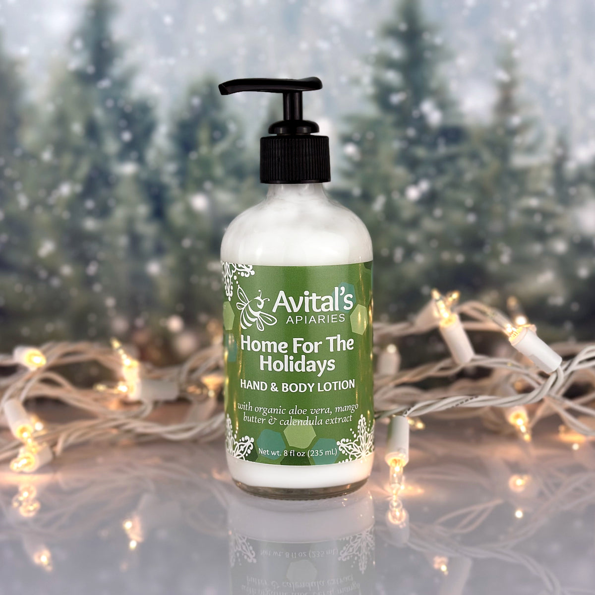 A bottle of Avital's Apiaries Hand & Body Lotion labeled 'Home For The Holidays' sits on a reflective surface with warm fairy lights and a blurred winter forest background.