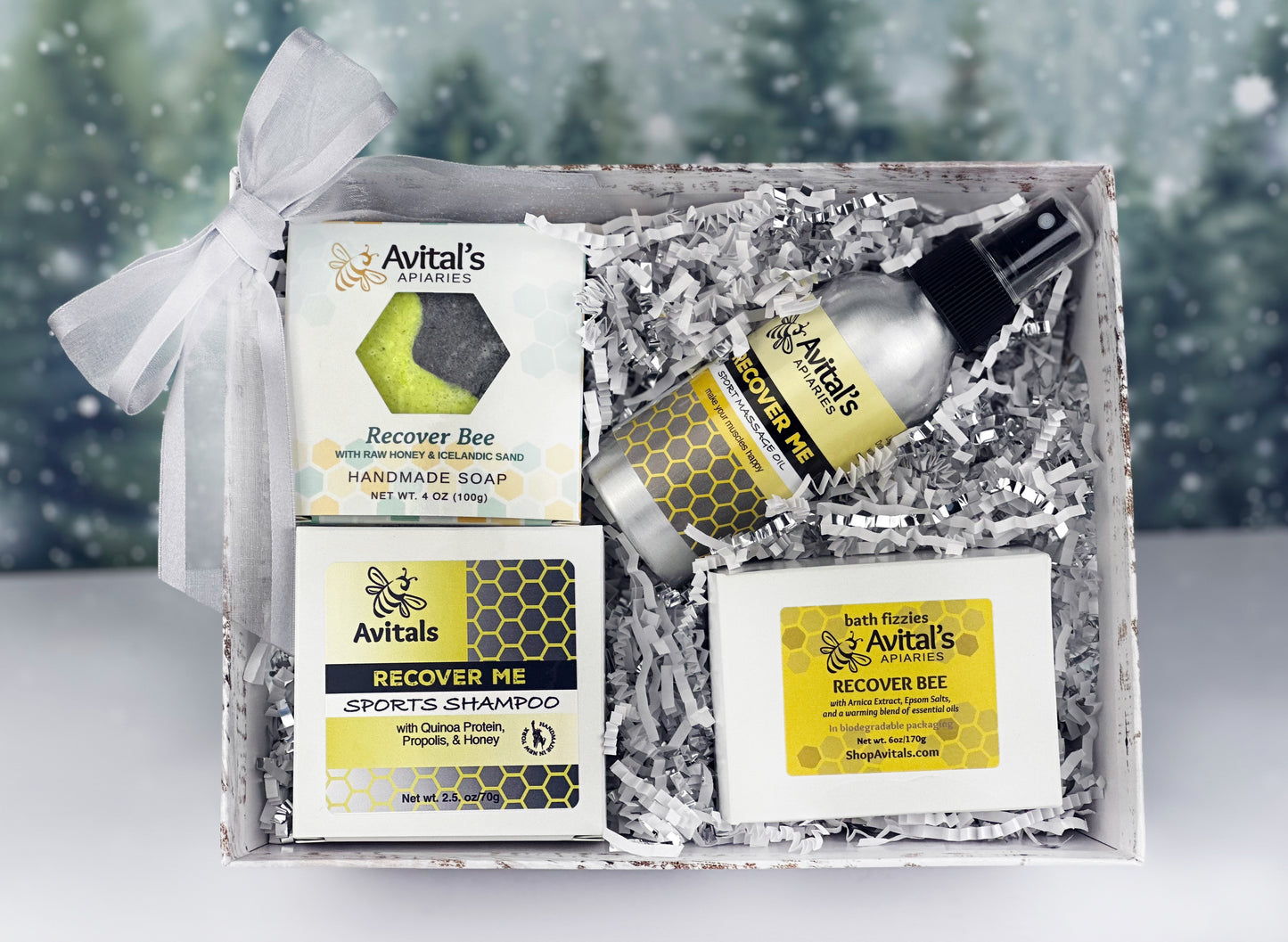 A gift set box containing Avital's APIARIES products, including Recover Bee soap, Recovery bath fizzies, Recover Me shampoo, and Recover Me body oil, with packaging that indicates they contain essential oils.