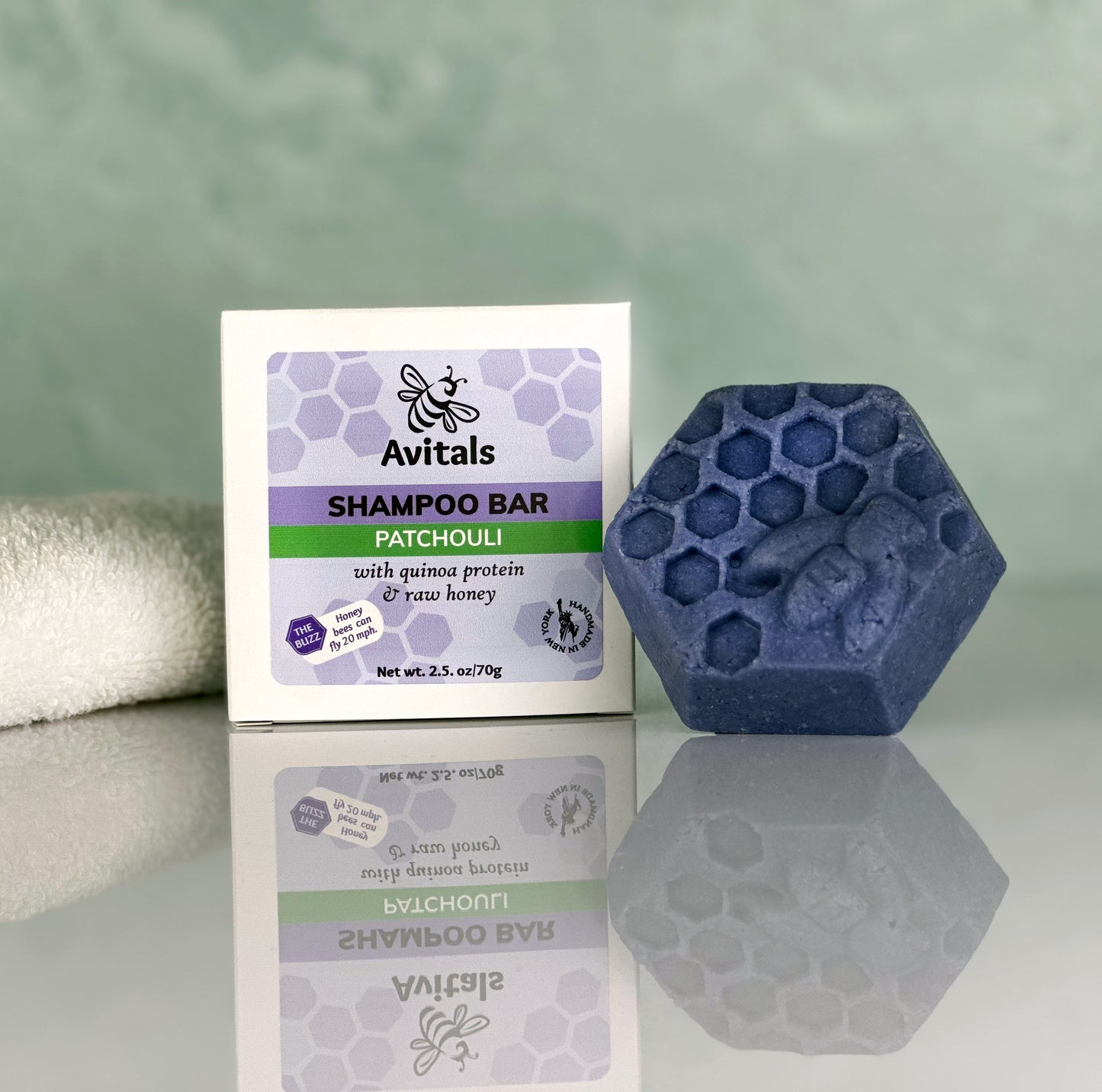 A Patchouli Shampoo Bar by Avitals, shown alongside its packaging. The bar is dark blue with a hexagonal design, placed on a white surface with a reflection, and the packaging is white with purple and green text and branding.