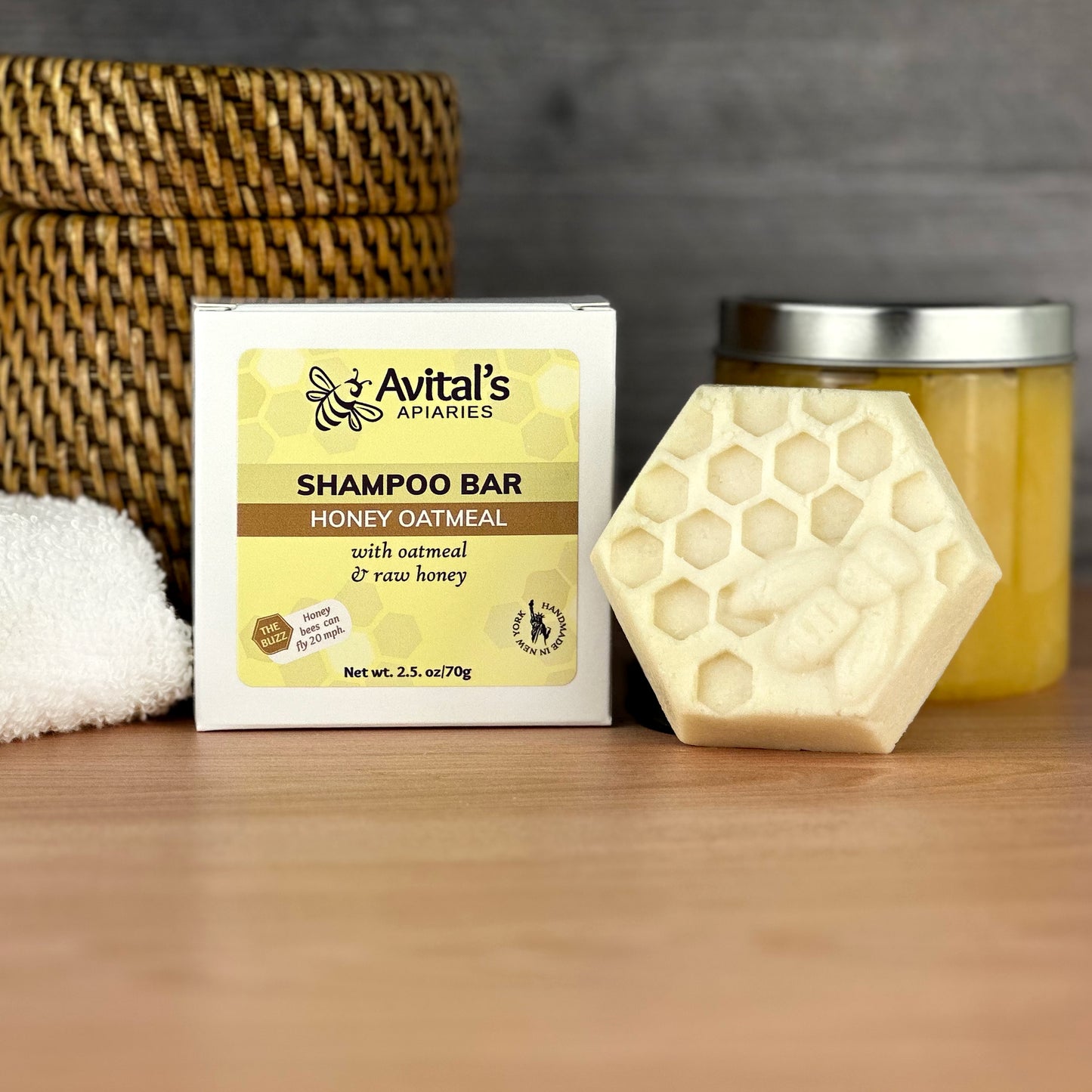 A honey oatmeal shampoo bar with its packaging, labeled 'Avital's Apiaries', displayed next to a jar of honey and a towel on a wooden surface.
