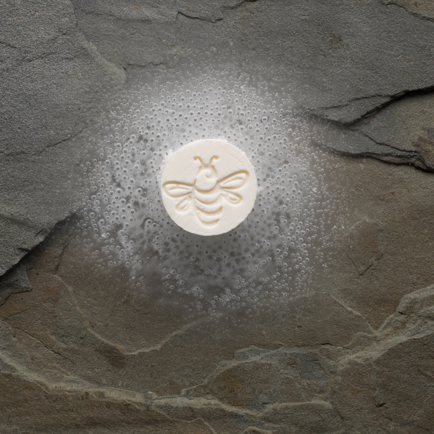 A beige shower steamer with a design resembling a bee in the center, placed on a dark, textured surface.