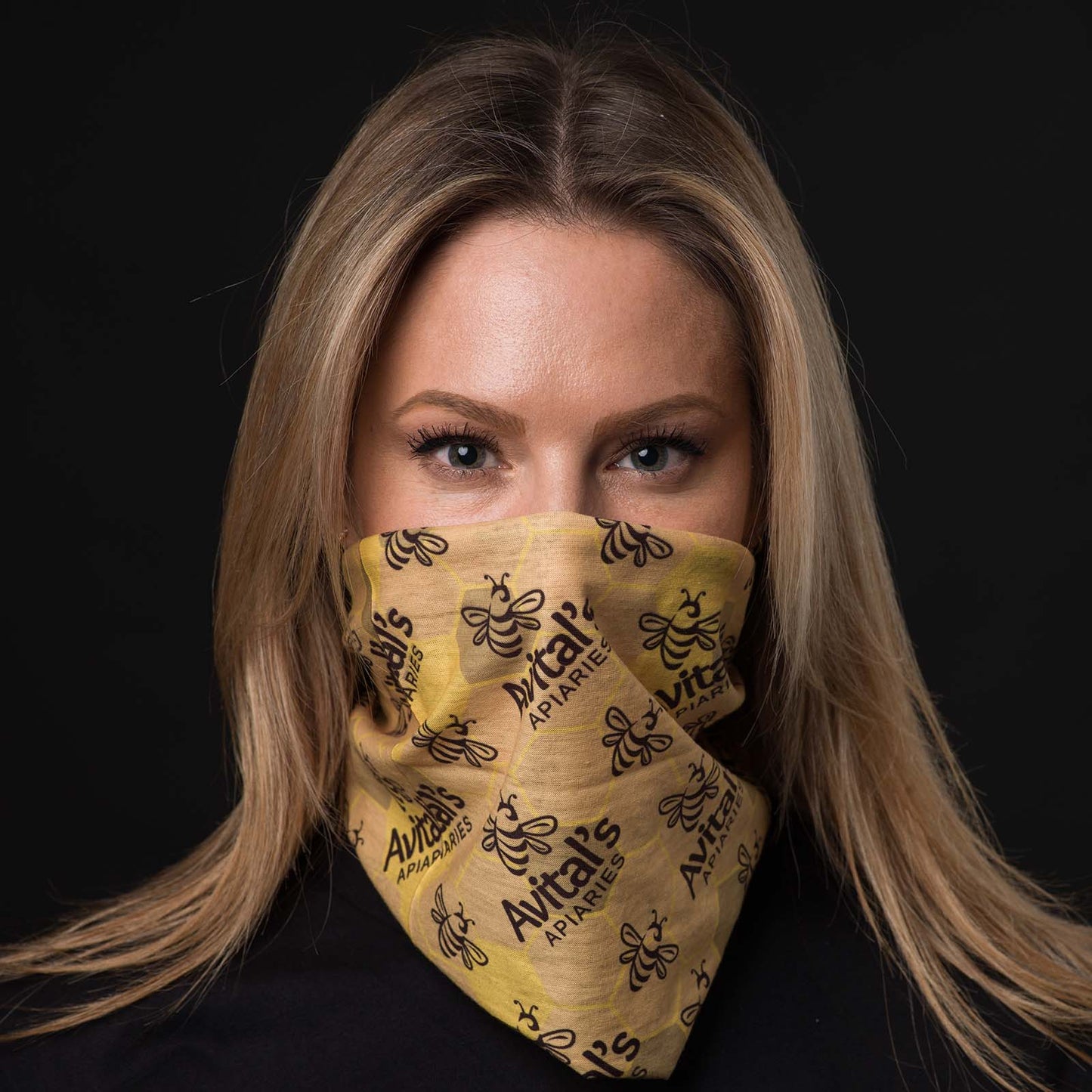 A blond woman smiling with an Avital's  tubular bandana pulled up over her nose, bandit style. The bandana is yellow with a dark brown Avital's logo and honey bee stamped on it.