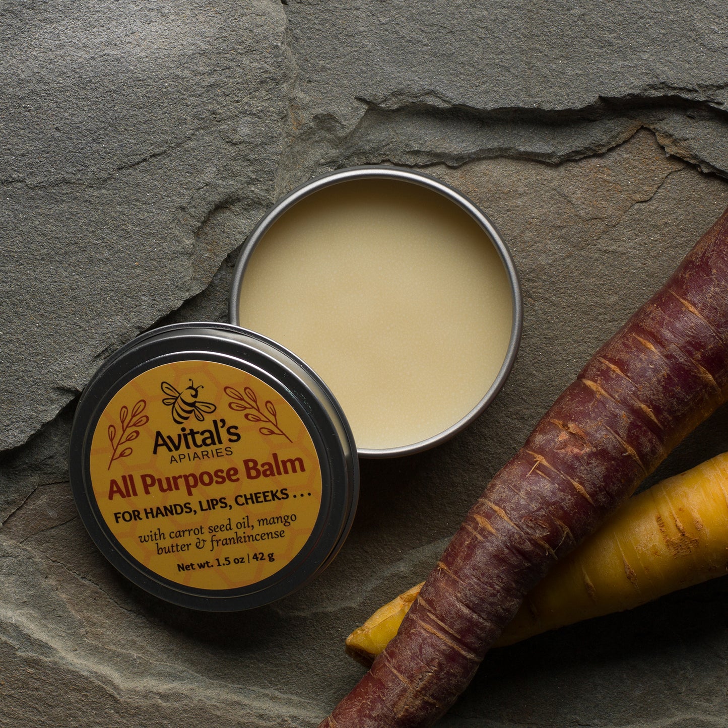 A tin of All Purpose balm open to show the light yellow balm. The label is yellow with red accents. Two carrots, one red, one yellow, are nearby.