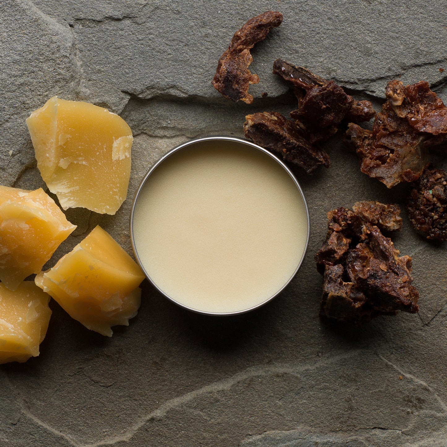 An open tin of Beekeeper's Buddy surrounded by chunks of beeswax and propolis on a gray slate background