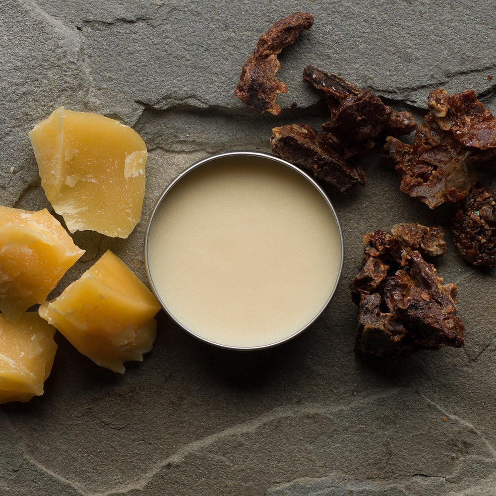 
                  
                    An open tin of Beekeeper's Buddy surrounded by chunks of beeswax and propolis on a gray slate background
                  
                