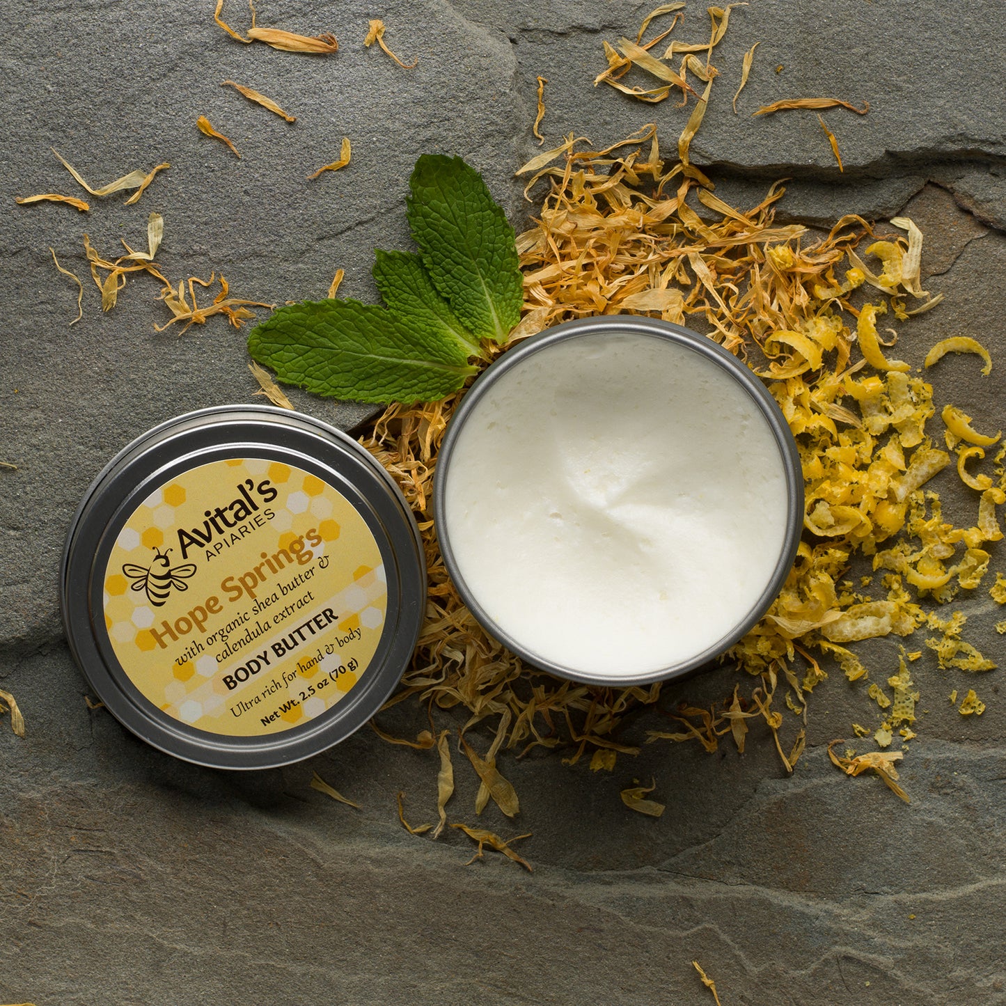 A tin container with a label reading 'Hope Springs Organic Body Butter' is open, showing the white solid contents. It is placed on a stone surface with scattered dried herbs and a mint leaf around it.