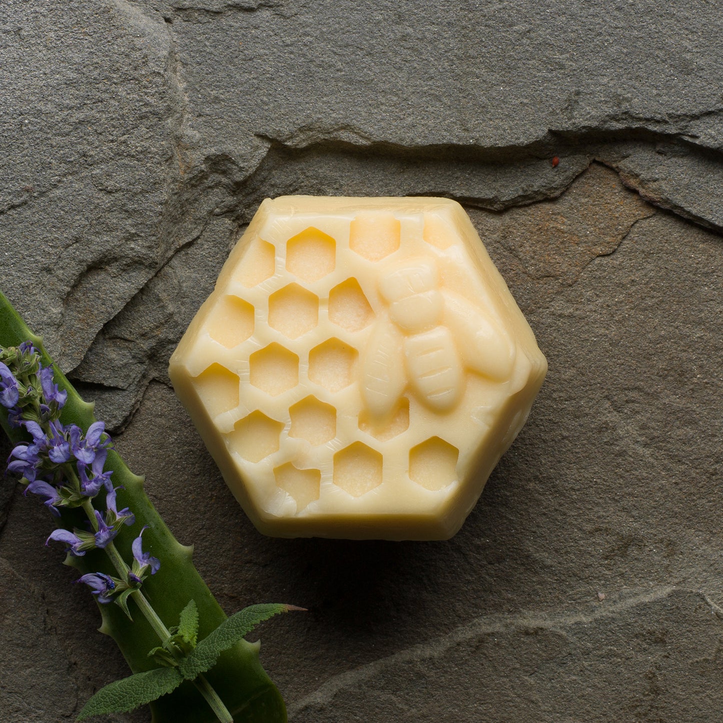 A cream-colored, hexagonal bar with a honeycomb and honey bee design. To the left is a sprig of lavender and an aloe leaf.