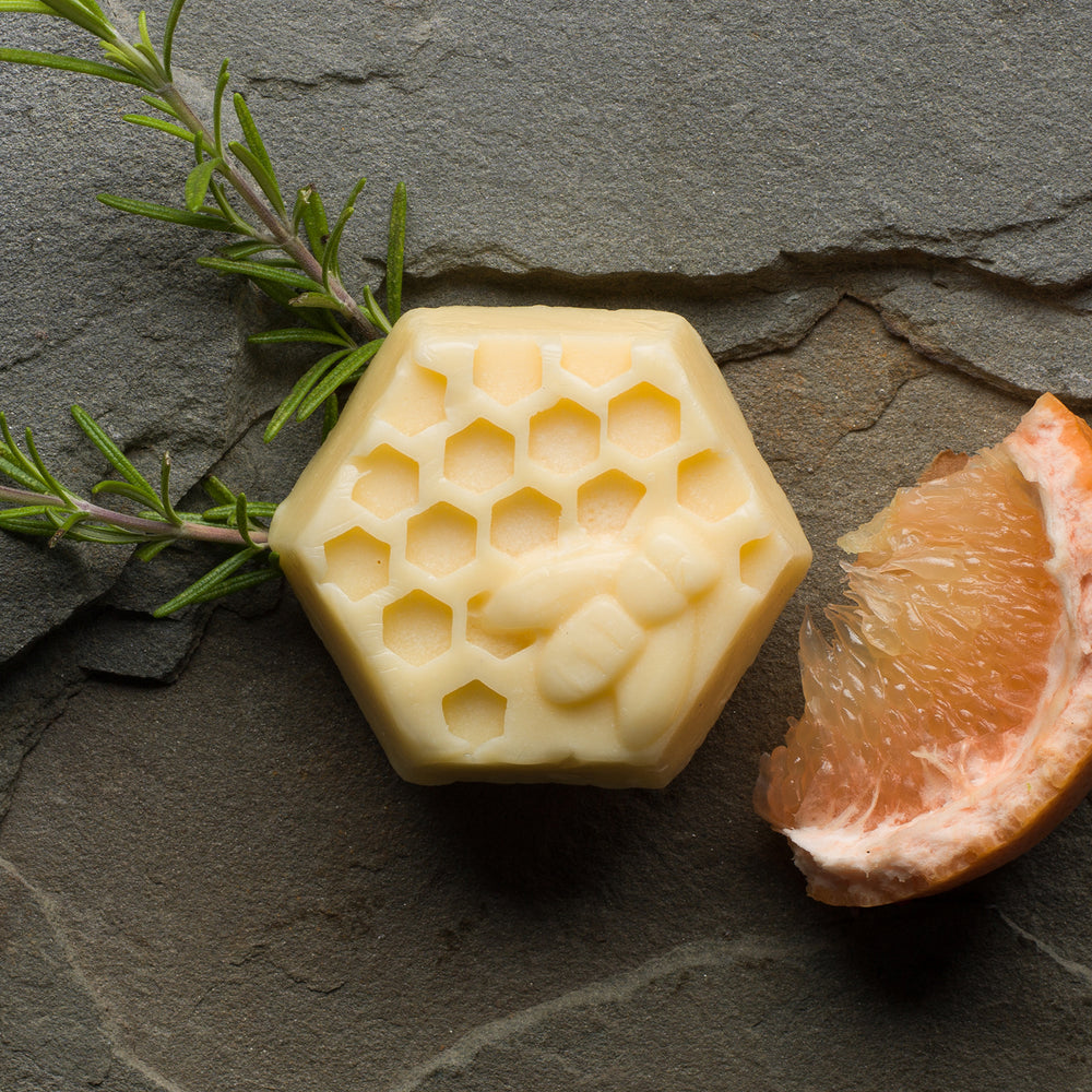 A cream-colored, hexagonal bar with a honeycomb and honey bee design. To the left is a a sprig of rosemary and a slice of bright yellow grapefruit
