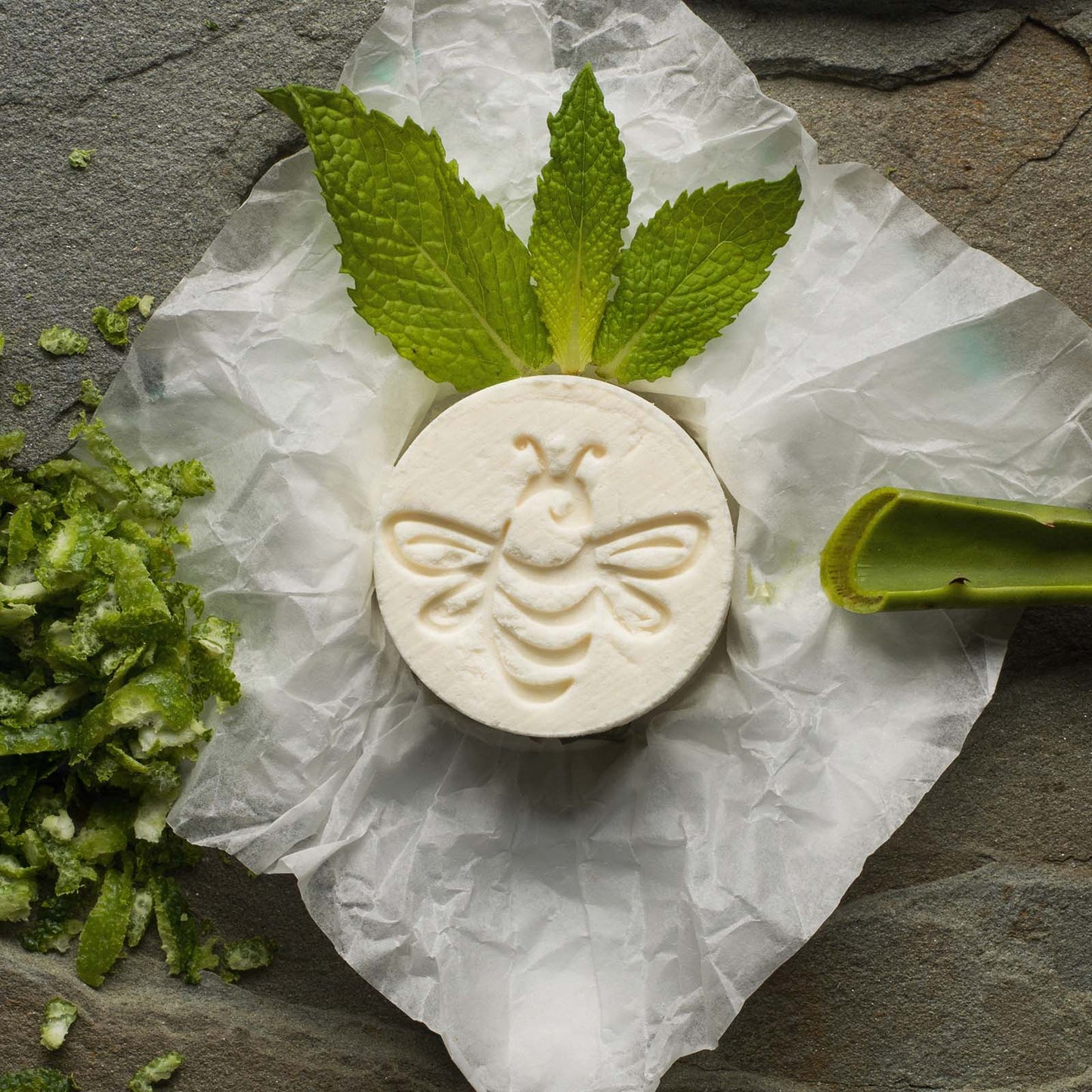 
                  
                    A cylindrical bee-stamped fizzy surrounded by bergamot peel, green spearmint leaves, and aloe 
                  
                