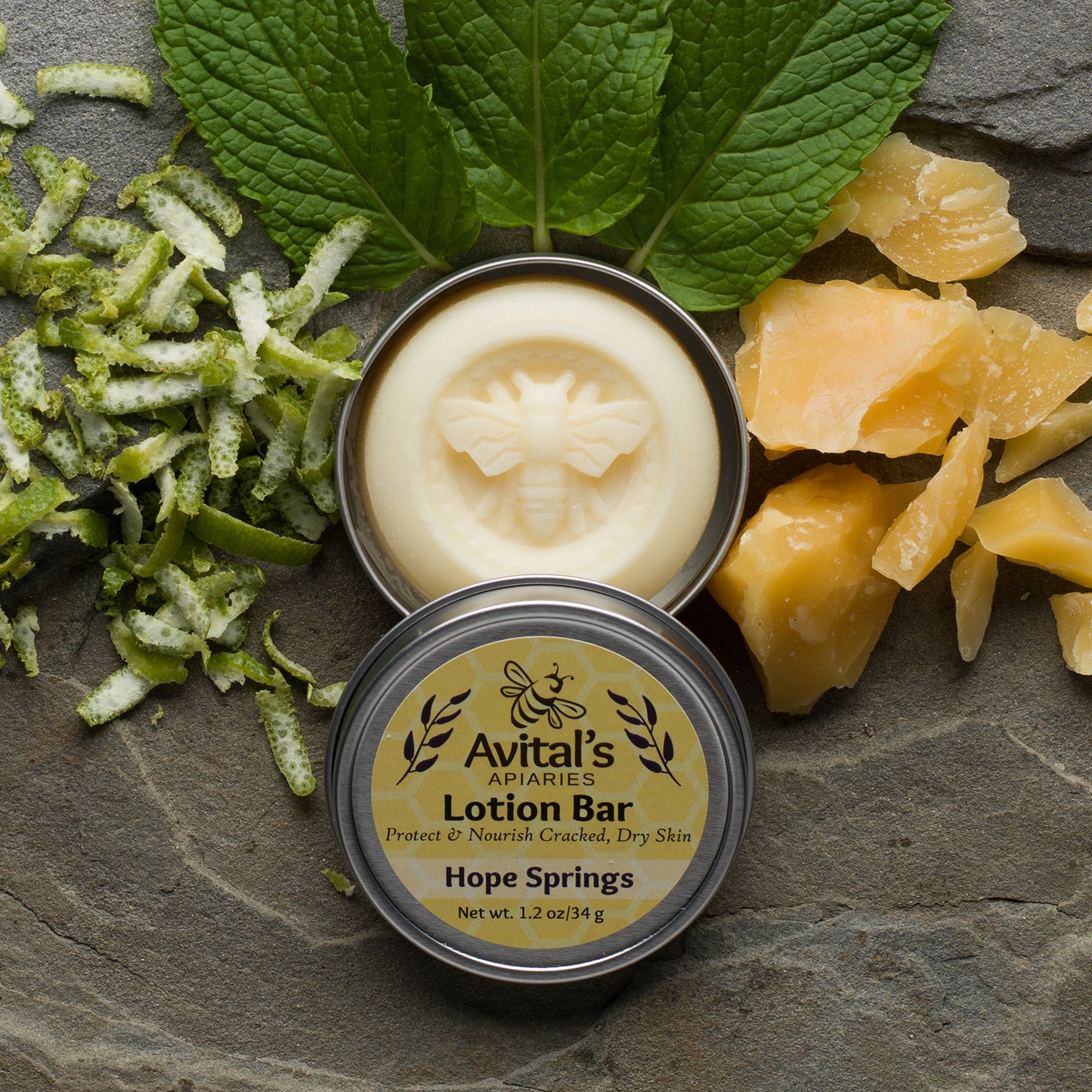 Overhead shot of a bee-stamped lotion bar in a tin. Around it is citrus peel, beeswax, and spearmint leaves. The label says "Protect & Nourish Cracked, Dry Skin."