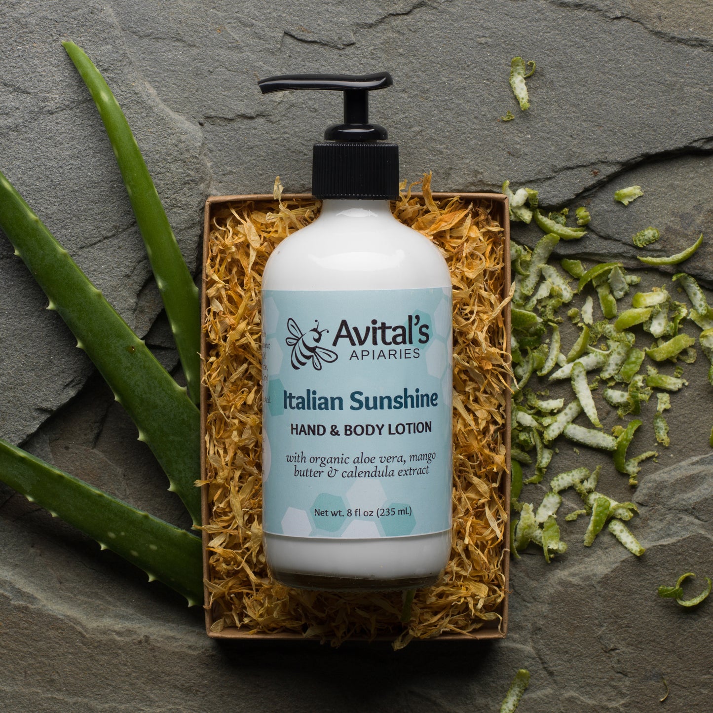 A white pump bottle labeled 'Avital's Apothecary Italian Sunshine Hand & Body Lotion' with green aloe leaves and a slice of mango in the background, presented on a grey stone surface.