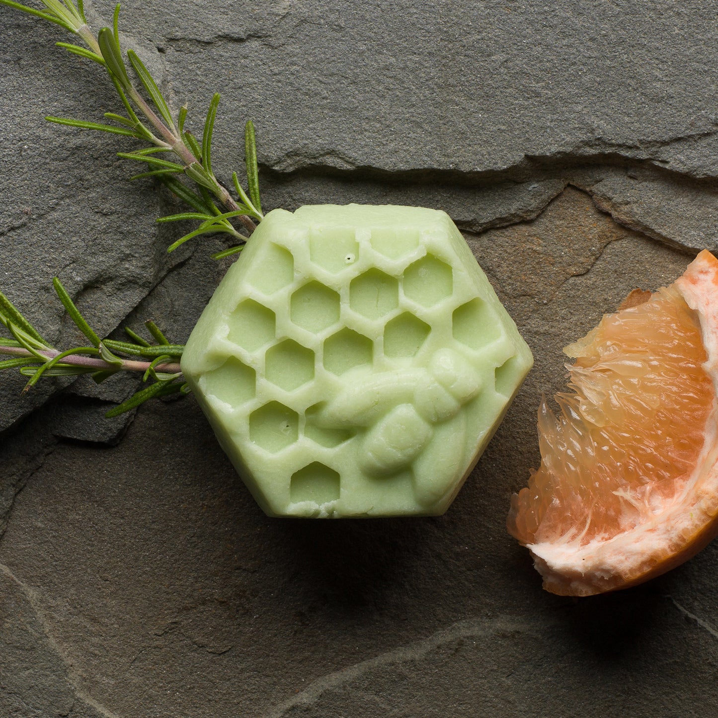 Hexagonal green bar with a honey bee and honeycomb on it. Next to it is a sprig of rosemary and a crescent of juicy grapefruit.