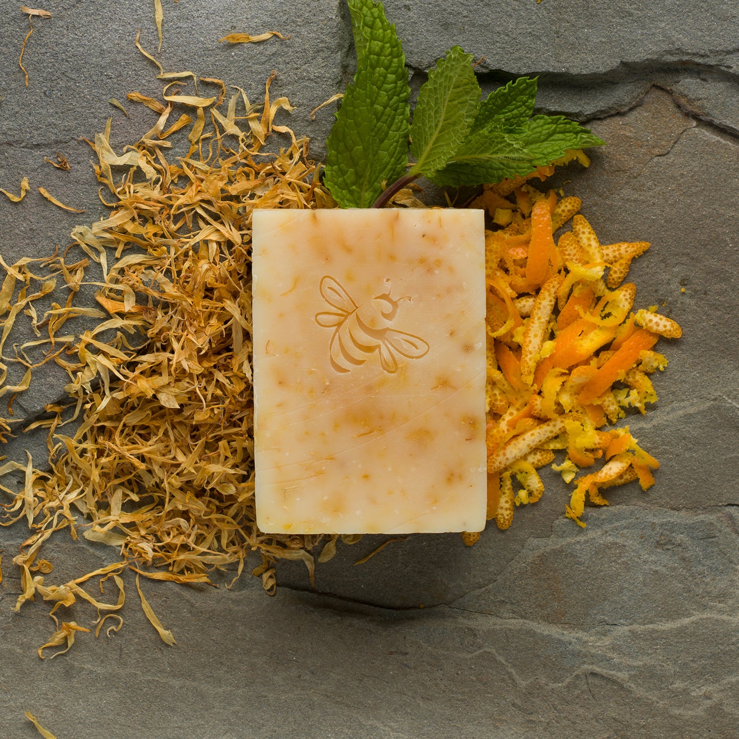 A yellow-orange, bee-stamped bar of Hope Springs soap surrounded by bright orange calendula petals, citrus peel shavings, and spearmint leaves on a dark gray slate.
