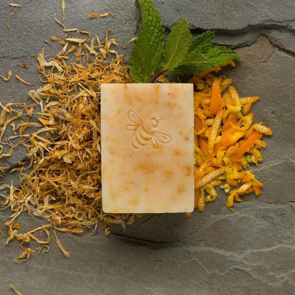 A yellow-orange, bee-stamped bar of Chaya's Hope surrounded by bright orange calendula petals, citrus peel shavings, and spearmint leaves on a dark gray slate.