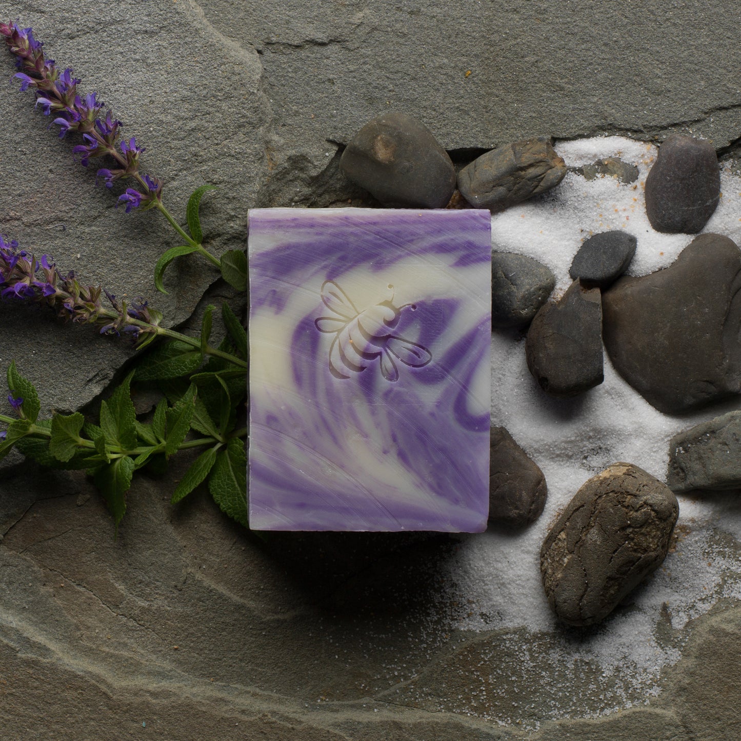 A bar of lavender and honey soap with a purple swirl, placed on a rock with lavender flowers and additional rocks around it.