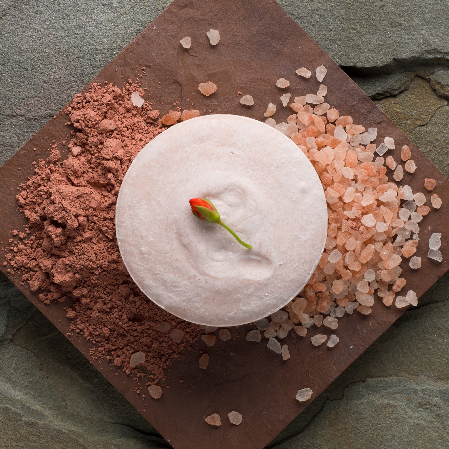 A pink bar of soap surrounded by pink salt with a geranium bud on top.
