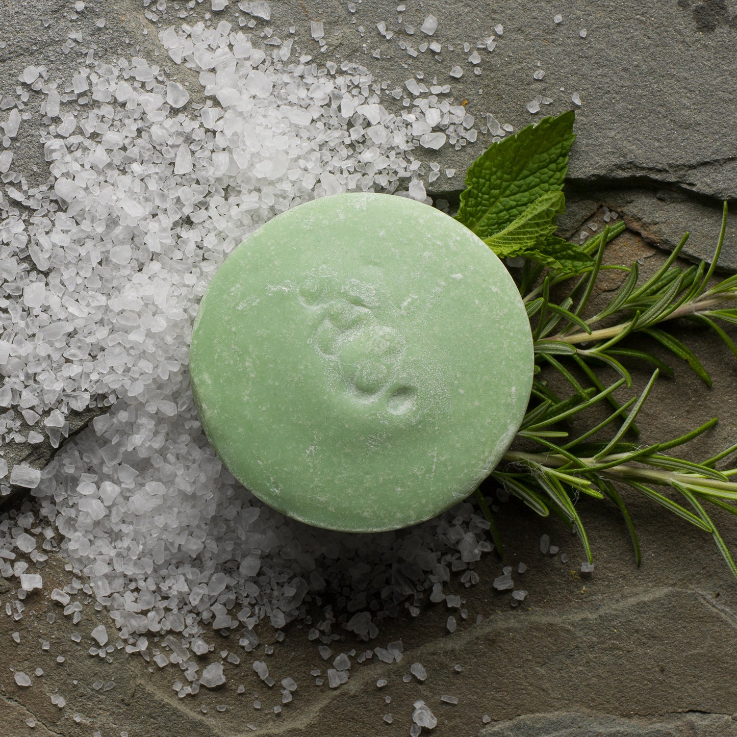 A round bar of soap with a green hue, placed on a surface with scattered white sea salt and adorned with a sprig of rosemary to the side.