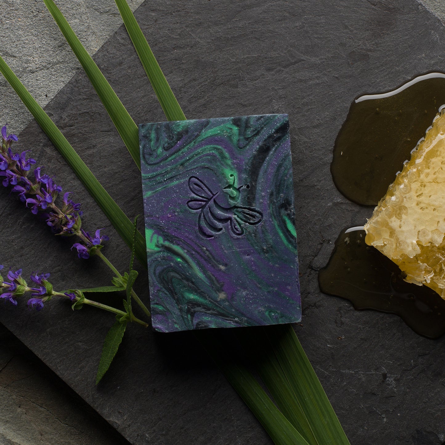 A bar of soap with swirled colors of purple and green, resembling the Northern Lights, placed on a dark surface with a piece of honeycomb and lavender flowers in the background.
